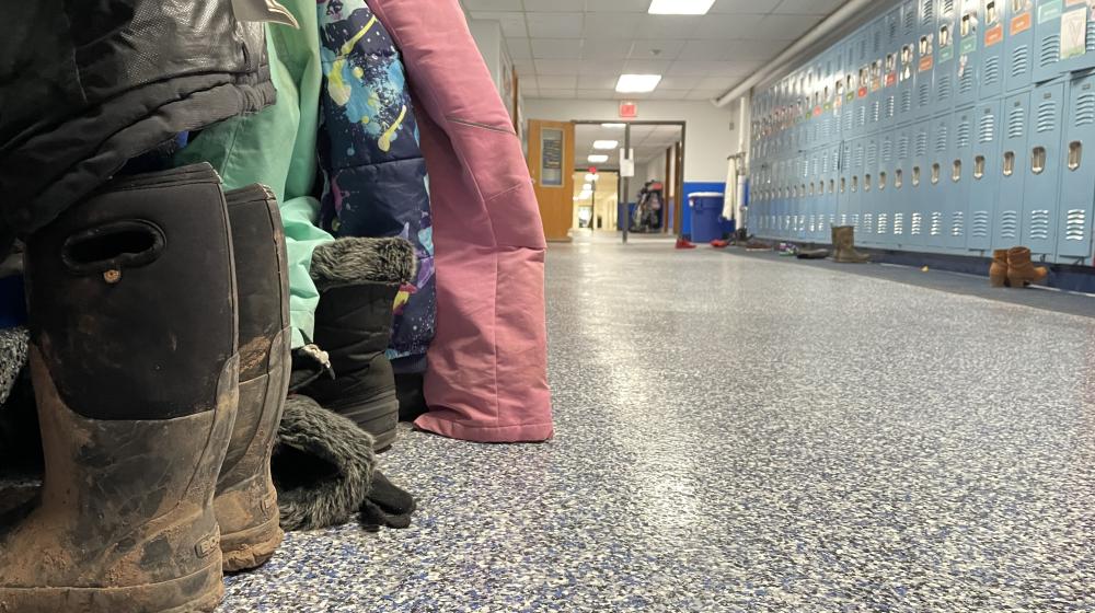 A hallway at a rural elementary school shows snowpants and boots across from a wall of blue lockers.