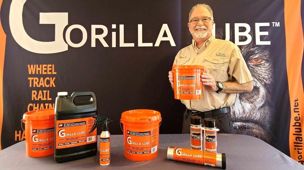 A bearded man poses in front of a backdrop that says "GORiLLA LUBE" with industrial products on a table in front of him with orange labels. 