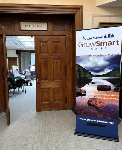 A banner with the GrowSmart Maine logo is set up outside the door to a  conference room at the Bangor Public Library. People sit at tables in the background, seen through the open wooden door.