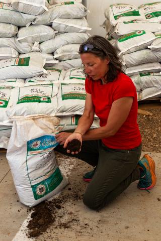 A woman kneels over an open bag of compost and holds a handful of brown soil-looking compost in her hand.