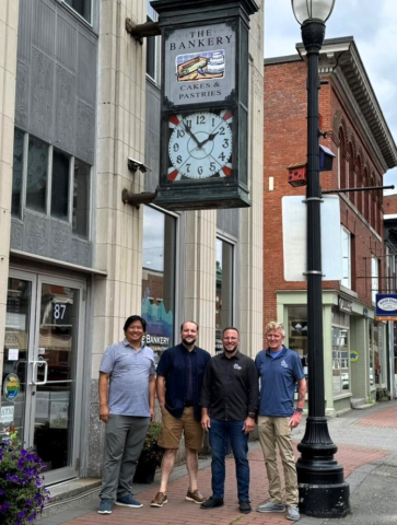 Four men stand on the sidewalk in front of The Bankery Fleuriste in Skowhegan. Over their heads is the vintage bank clock that is part of the business's sign. Brick buildings can be seen in the background. 