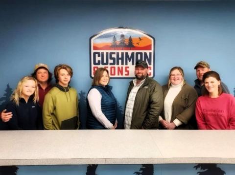 Eight people of various ages stand in front of a sign that reads "Cushman and Sons Inc."