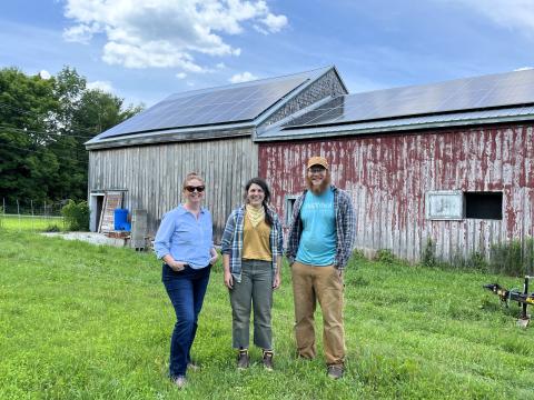 Three people stand in front of a barn with solar panels covering the roof. There is a lawn in front and trees behind.