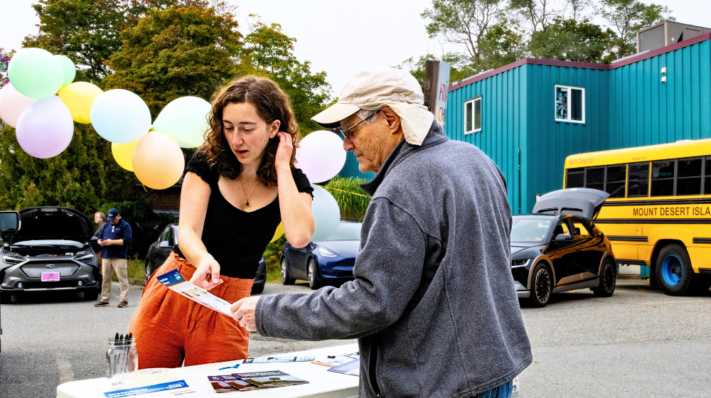 The Climate Fellow described in the story is pictured at an outdoor table interacting with a community member who is reading a pamphlet she shows him. There is a blue building, a Mount Desert Island School bus, and trees in the background. The event pictured is in a parking lot, with balloons in the near background. 