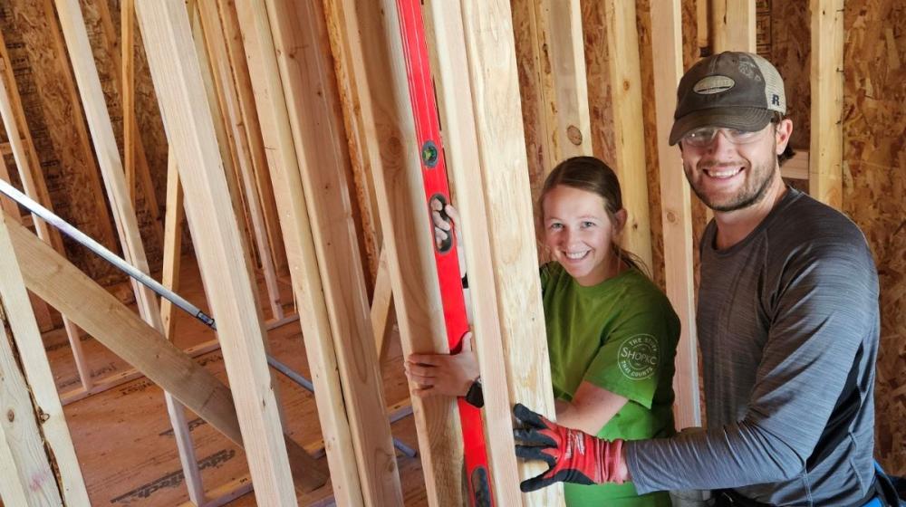 Cody (Caucasian white male wearing black and grey jacket with matching ballcap) and Melissa (Caucasian young adult female wearing olive green shirt)Olsen in Hyrum, Utah in June of 2024, seen standing within the wooden framework of their Mutual Self-Help house.