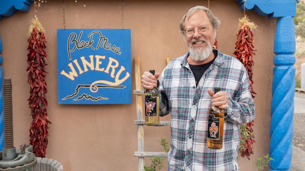 Man holding two bottles of wine in front of Black Mesa Winery sign