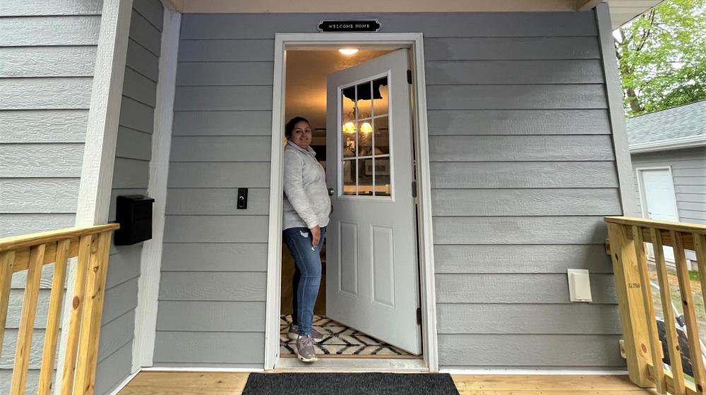 Smiling woman standing in the open doorway of her home as seen from the porch of the home.