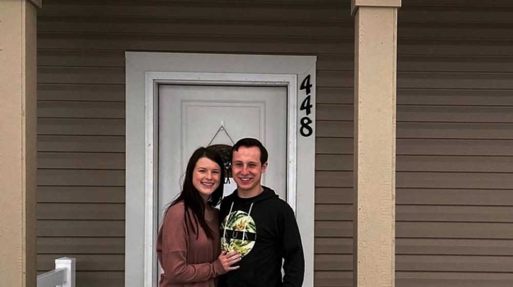 Caucasian couple with light skin, dark hair, stand on porch of their new house with brown siding and tan brick.