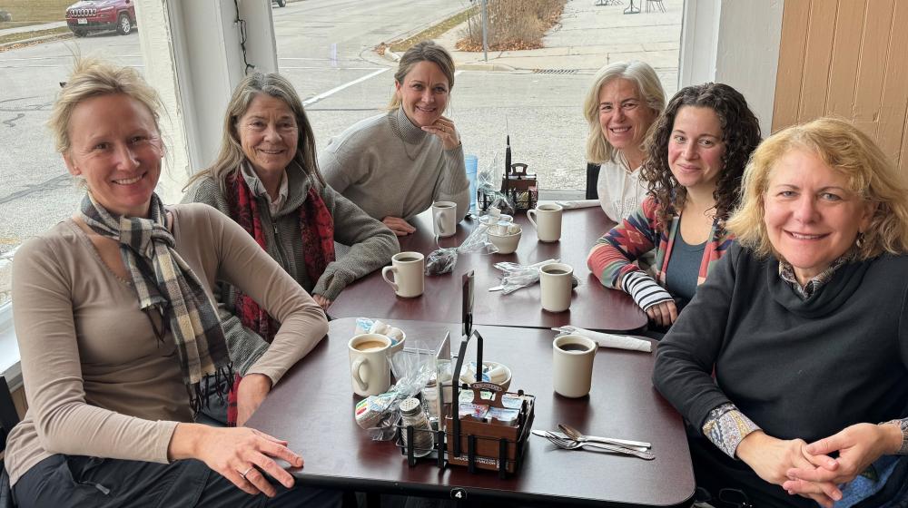 Six women sit around a table drinking coffee and tea