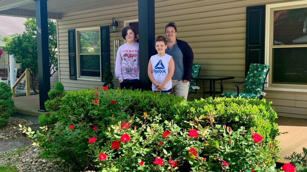 Mom and two sons standing on the porch of their two-story house, as seen from the yard,