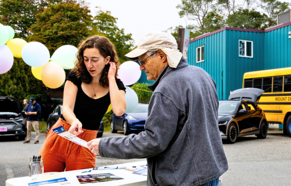 The Climate Fellow described in the story is pictured at an outdoor table interacting with a community member who is reading a pamphlet she shows him. There is a blue building, a Mount Desert Island School bus, and trees in the background. The event pictured is in a parking lot, with balloons in the near background. 